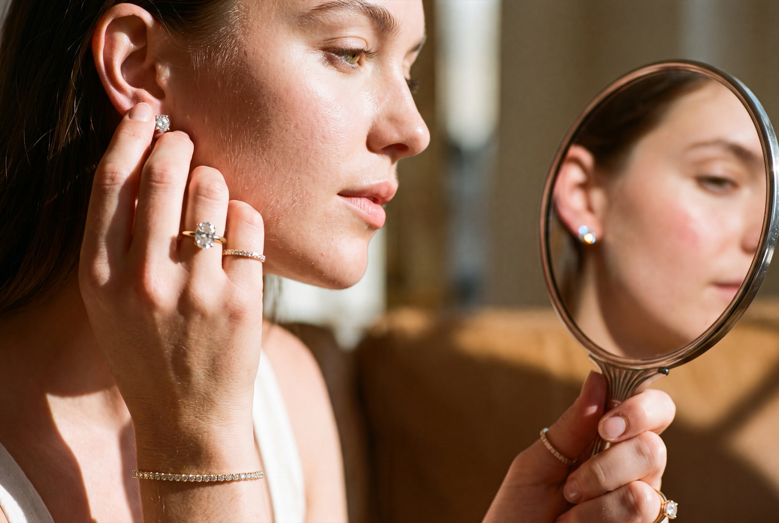 Woman admiring a lab-grown diamond stud earring in a hand mirror, wearing rings and a tennis bracelet